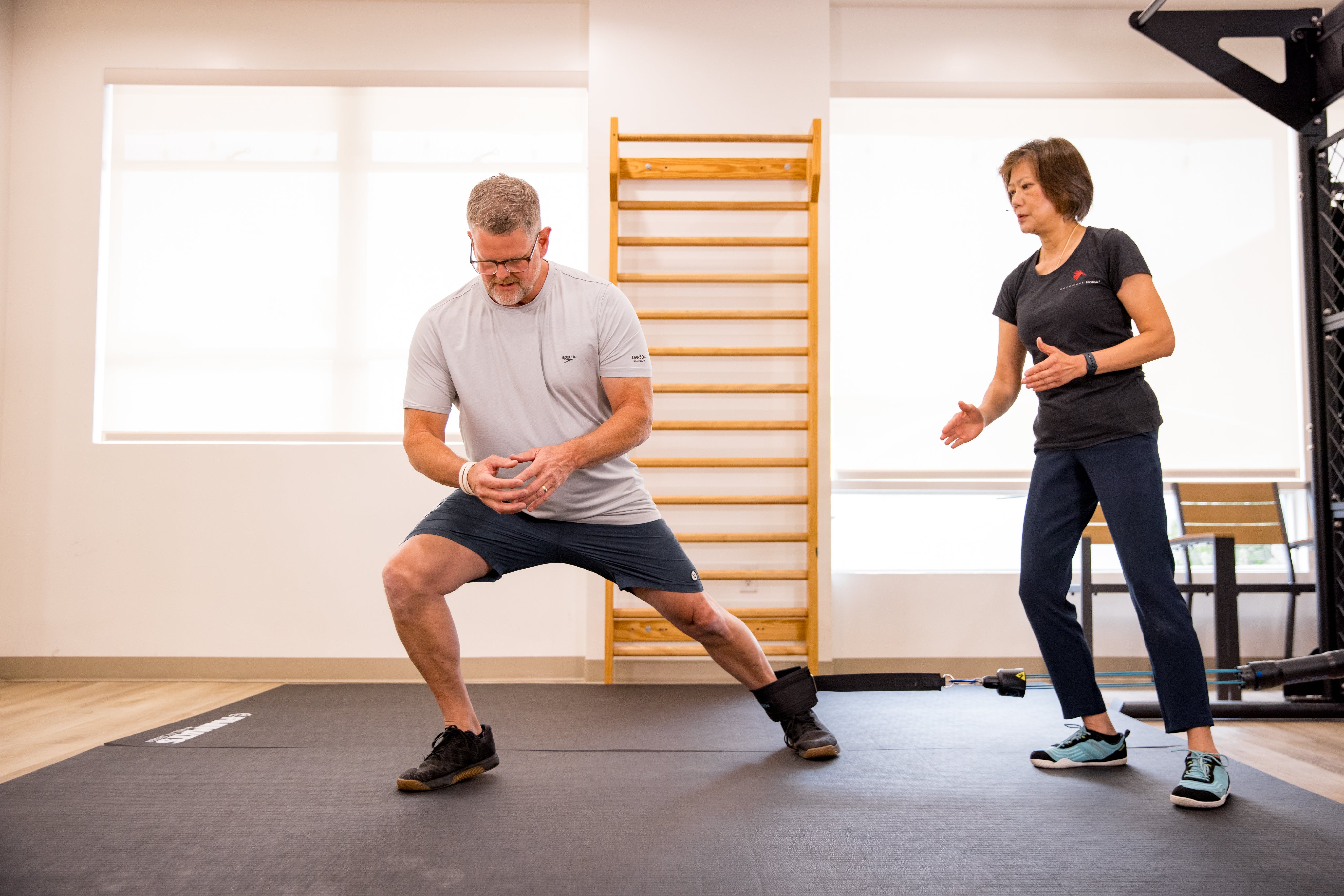 Man performing a lateral lunge exercise with a resistance band attached to his ankle, while a trainer watches and provides guidance